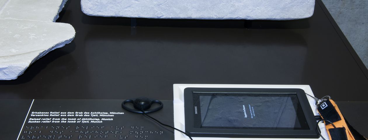 Auf einem Tisch vor dem Objekt links ein Austellertext in Braille-Schrift und rechts ein Tablet- Ausstellerguide.