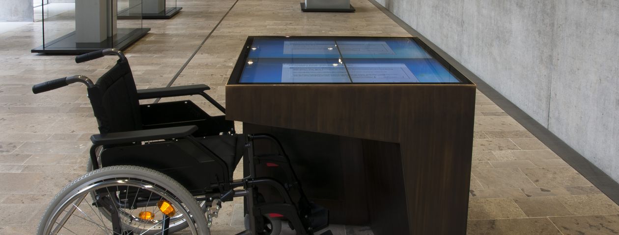 Ein Rollstuhl vor einem unterfahrbaren ScreenTouch-Tisch Auf dem Boden das taktile Leitsystem, im Hintergrund einzelne Steinobjekte auf Blöcken. 