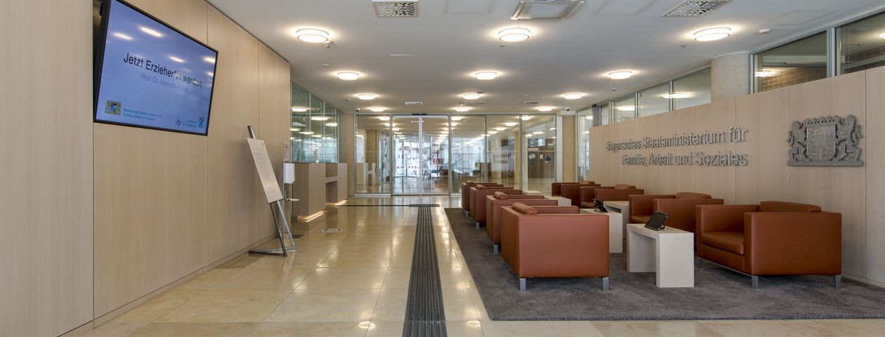 Blick in das Foyer vom Eingang aus. Dunkles taktiles Leitsystem im hellen Marmorboden Rechts eine Anordnung von Sitzelementen, links an der Wand ein Bildschirm