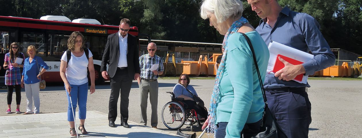 Im Vordergrund eine Person mit Blindenstock vor dem taktilen Leitsystem mit einem Begleiter. Im Hintergrund mehrere Personen, unter anderem eine Rollstuhlfahrerin. 