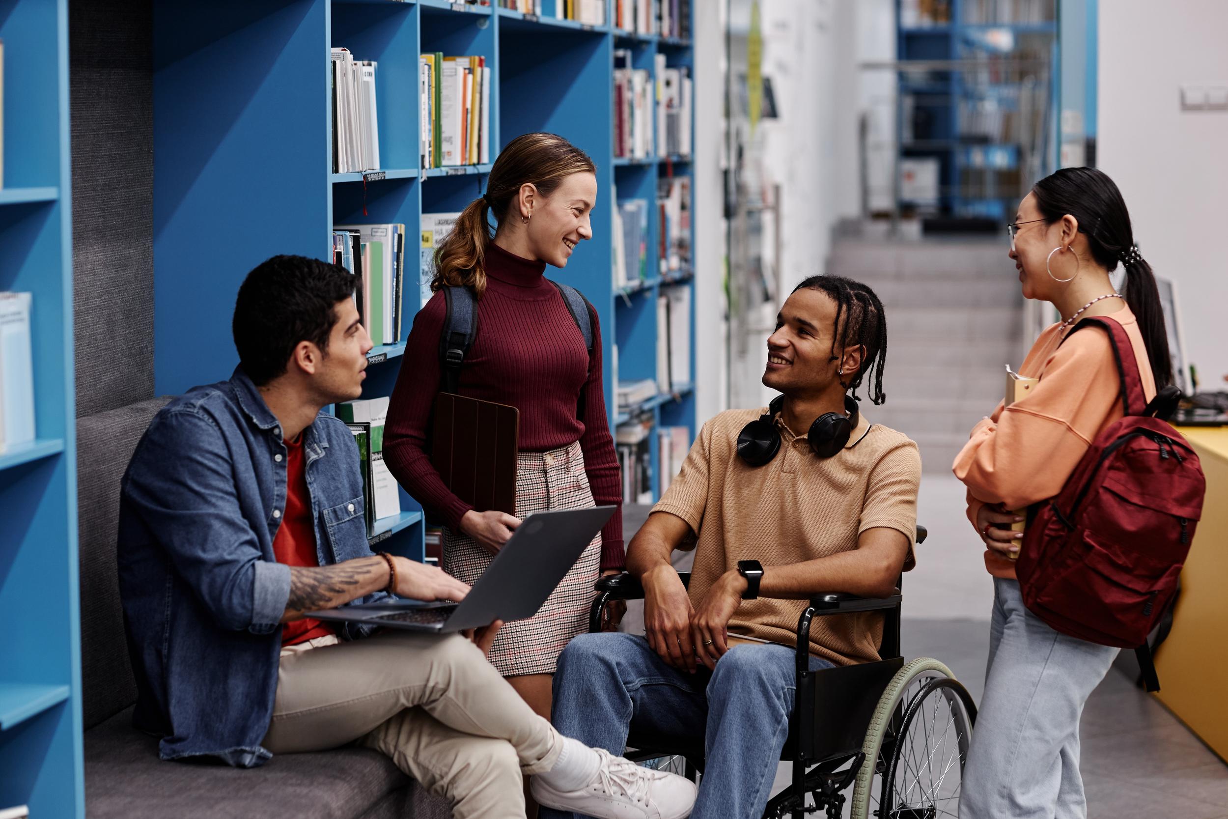 Eine Gruppe von jungen Studentinnen und Studenten, darunter ein Mann im Rollstuhl, unterhalten sich fröhlich in einer Bibliothek.