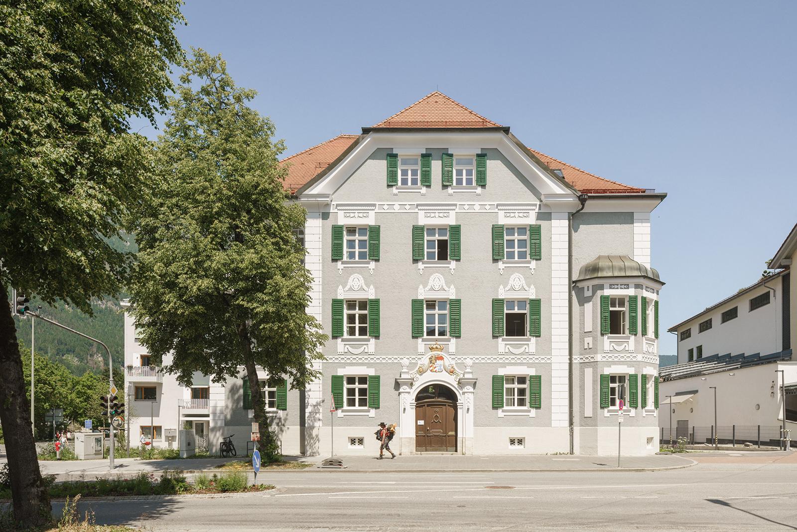 Straßenansicht des viergeschossigen Gebäudes aus der Jahrhundertwende mit hellgrauer Fassade, grünen Holzfensterläden, weiße Dekorationselementen und einem prunkvoll sanierten Eingangsportal.