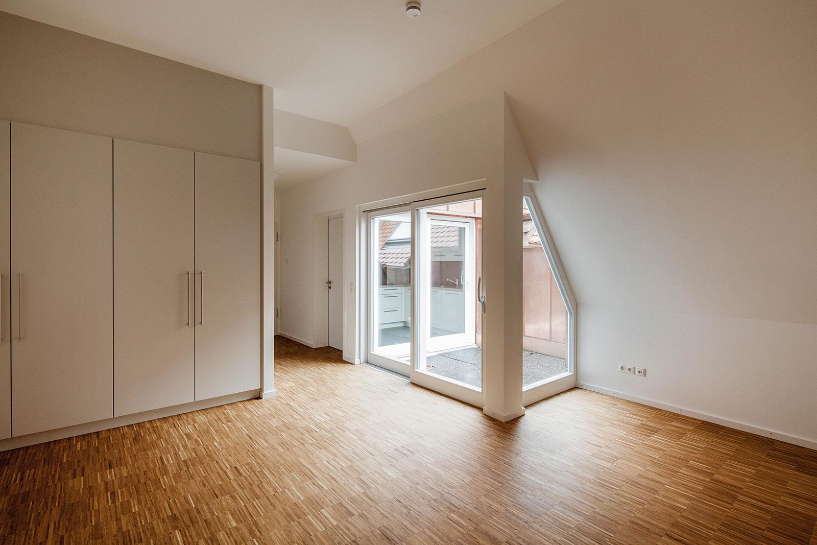 Ein leeres Schlaf- oder Wohnzimmer mit Holzfußboden und weißen Einbauschränken sowie einer kleinen angeschlossenen Loggia.