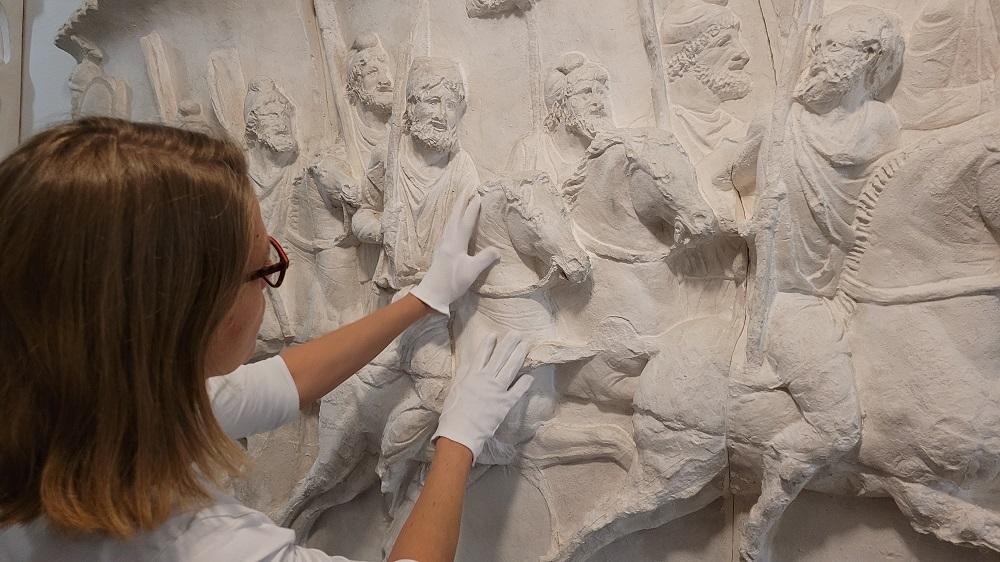 Eine Frau mit weißen Handschuhen ertastet die Details eines Wandreliefs.