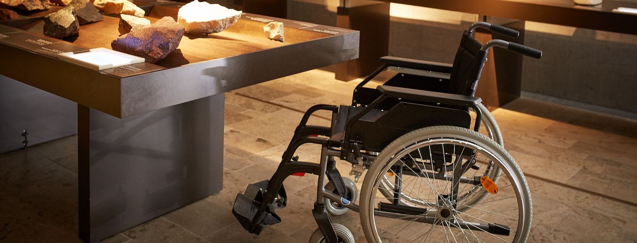 Ein Tisch mit Steinobjekten zum Befühlen. Dank der geringen Höhe und Gestaltung ist der Tisch mit dem Rollstuhl, der davor steht, unterfahrbar. 