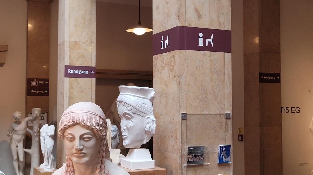 Ein braunes Farbband, angebracht an den Säulen und Wänden in etwa zwei Metern Höhe, weist mit weißer Beschriftung und Symbolen auf die verschiedenen Einrichtungen im Museum hin. Im Vordergrund befinden sich Gipsbüsten.