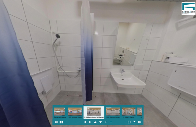 Der Screenshot des barrierefreien WCs zeigt links eine bodengleiche Dusche, daneben ein unterfahrbares Waschbecken, im Spiegel ist die behindertengerechte Toilette zu sehen.