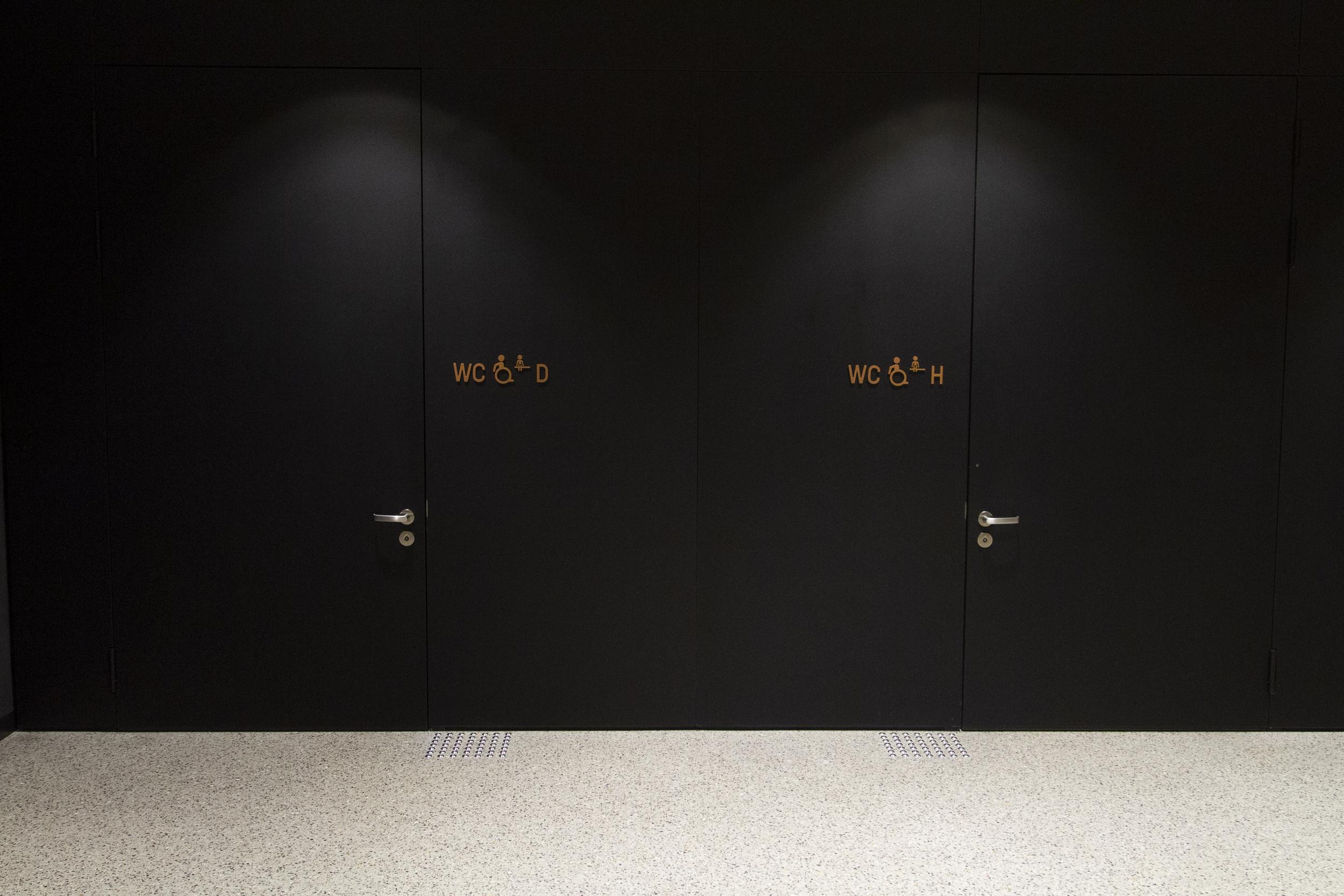 Im Hintergrund 2 Türen zu zwei rollstuhlgerechten Toiletten, links für Damen, rechts für Herren, mit jeweils einem Aufmerksamkeitsfeld (auf dem Boden) neben den Türen.  Die Türgriffhöhe ist barrierefrei.