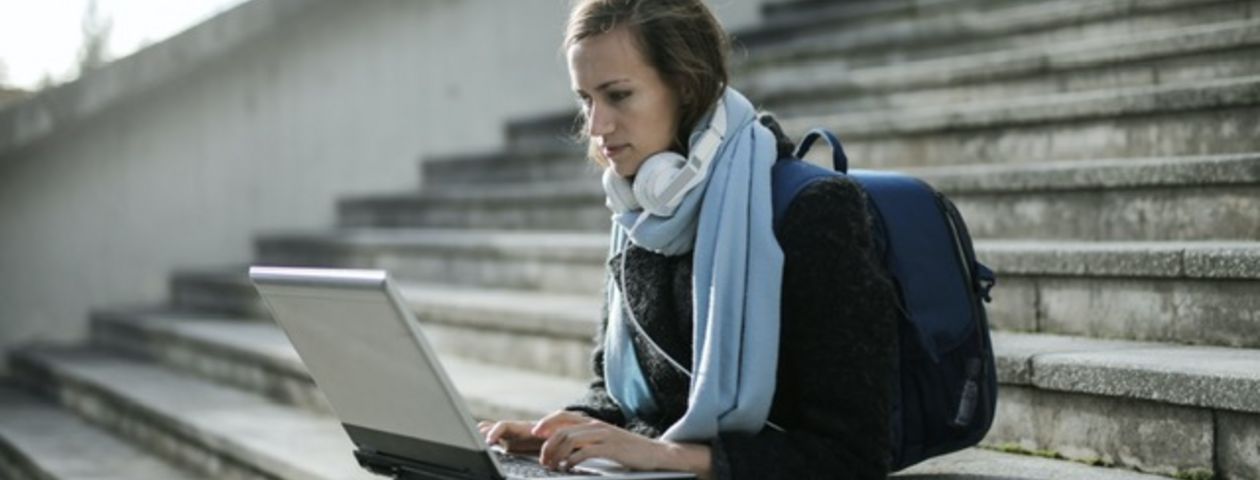Eine junge Frau mit Rucksack, Schal und Kopfhörern sitzt mit ihrem Laptop auf einer langen bzw. hohen Außentreppe. Sie schaut auf den Bildschirm und tippt etwas ein.