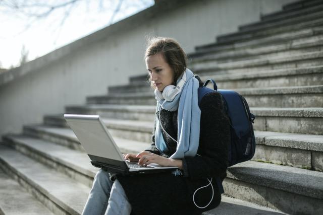 Eine junge Frau mit Rucksack, Schal und Kopfhörern sitzt mit ihrem Laptop auf einer langen bzw. hohen Außentreppe. Sie schaut auf den Bildschirm und tippt etwas ein.
