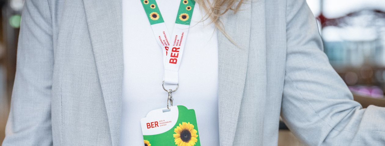Eine Frau mit schulterlangem Haar, weißem Shirt und grauem Jackett hat das Sunflower-Trageband umgehängt. Dieses ist in den Farben grün-weiß gehalten und mit Sonnenblumen und den roten BER-Schriftzügen des Flughafens gestaltet. Um sie herum sieht man verschwommen eine Flughafenatmosphäre.
