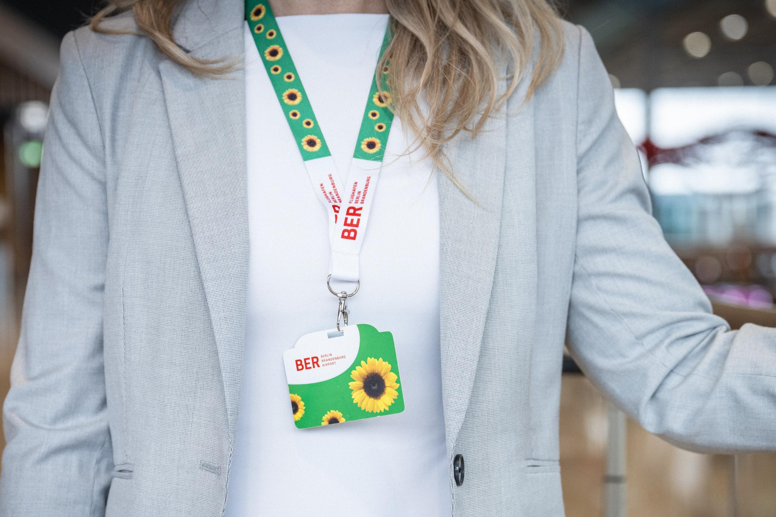 Eine Frau mit schulterlangem Haar, weißem Shirt und grauem Jackett hat das Sunflower-Trageband umgehängt. Dieses ist in den Farben grün-weiß gehalten und mit Sonnenblumen und den roten BER-Schriftzügen des Flughafens gestaltet. Um sie herum sieht man verschwommen eine Flughafenatmosphäre.