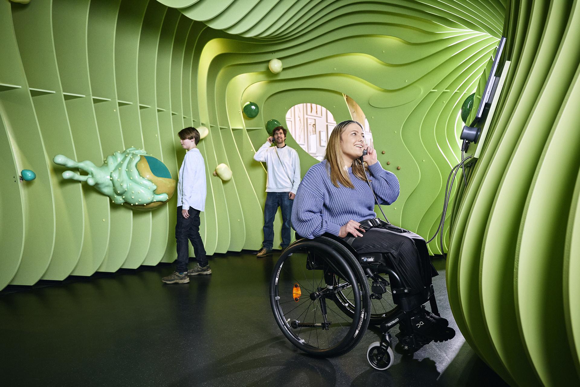 Drei Museumsbesucher, darunter eine Frau im Rollstuhl, hören Audioinformationen in einer überdimensionalen begehbaren Pflanzenzelle.