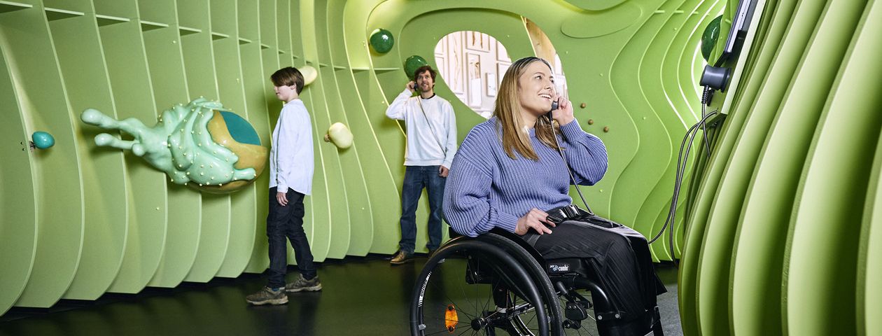 Drei Museumsbesucher, darunter eine Frau im Rollstuhl, hören Audioinformationen in einer überdimensionalen begehbaren Pflanzenzelle. 