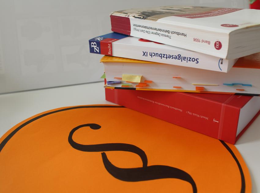 Rechts liegt ein Bücherstapel. Davor sieht man einen großen Pagraphen auf einem orangen Plakat.