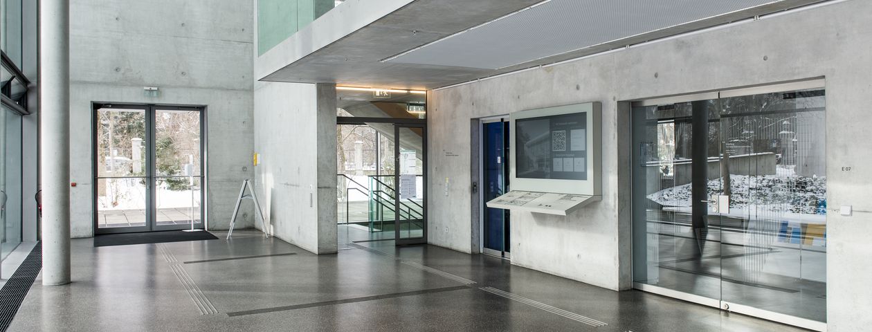 Innenansicht Eingangshalle auf den Haupteingang Ein textiles Leitsystem auf dem Boden führt zu den Treppen und  zu dem taktilen Lageplan an der rechten Wand  Links die verglaste Außenwand