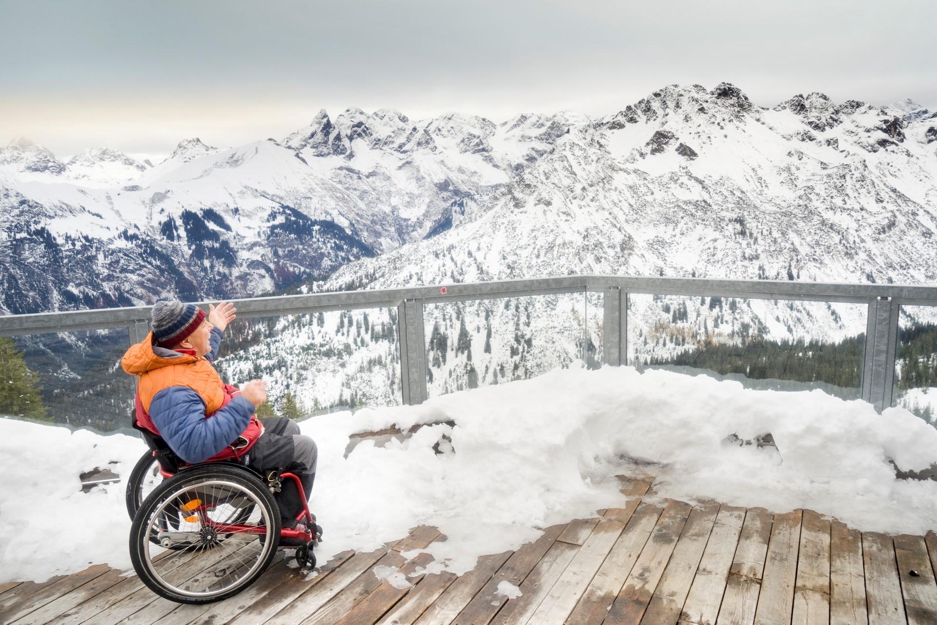 Eine Person im Rollstuhl blickt von einer Holzterrasse begeistert auf das verschneite Alpenpanorama.