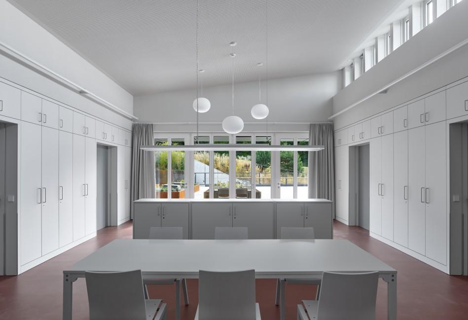 Blick durch den Wohnraum auf die Terrassen. Der Fußbodenbelag in einem dunklen rötlichen Farbton bildet einen Kontrast zu den hellen Schränken links und rechts und der grauen Möblierung wie etwa dem großen Tisch im Vordergrund.
